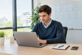 Young businessperson working on a laptop at a clean desk in a sunlit modern office, with a notepad, smartphone, and a whiteboard in the background.